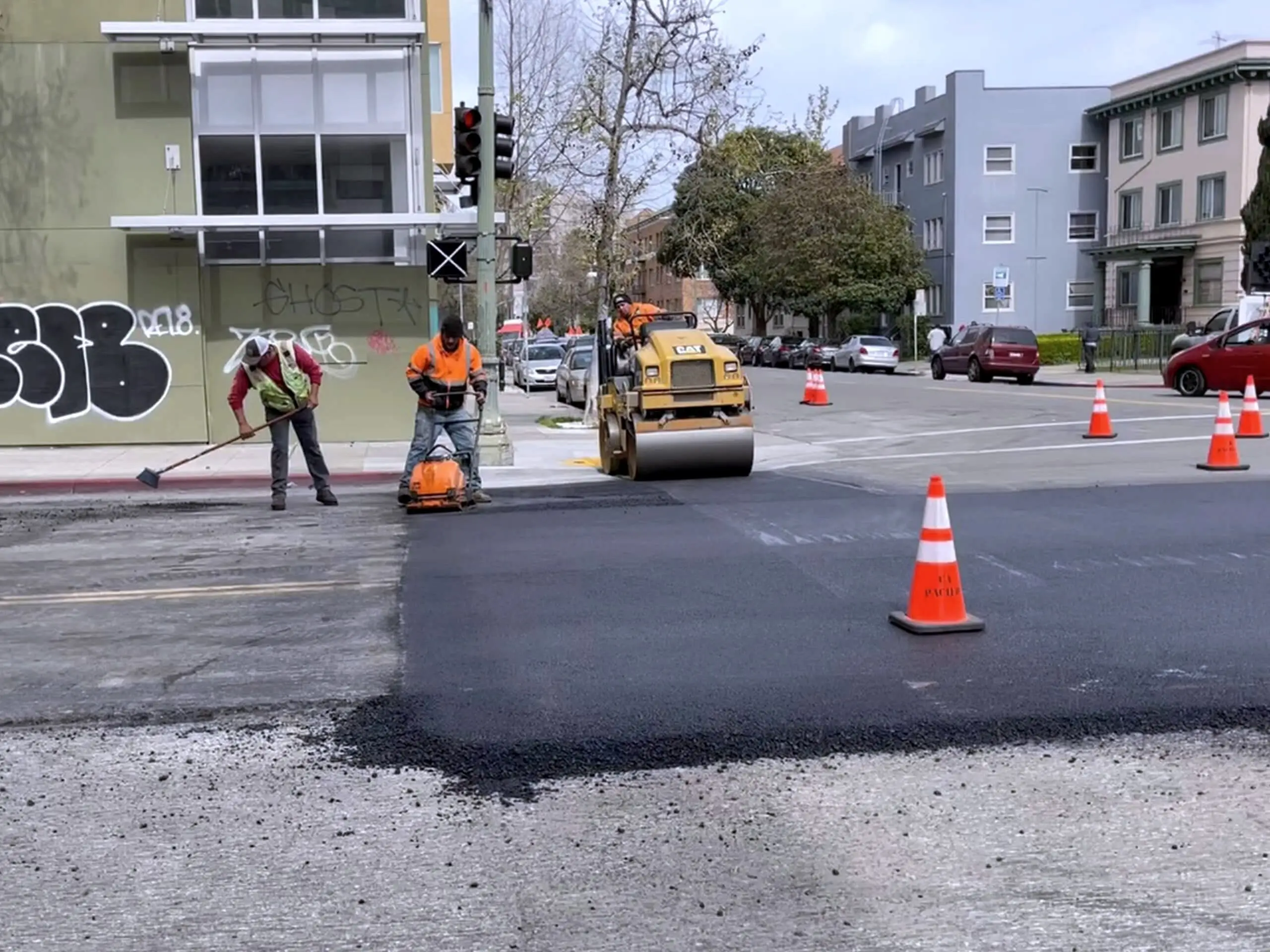 Two Forticon crew members working a roller across newly placed asphalt at a downtown Oakland intersection.