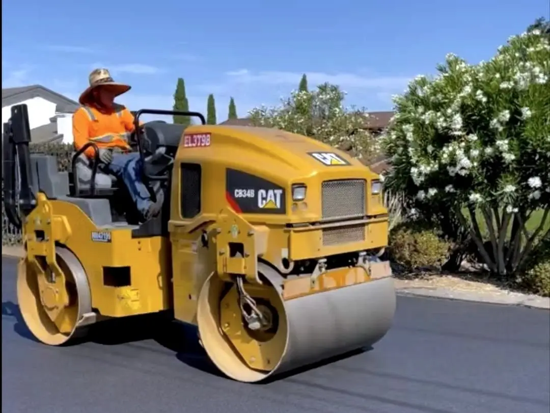 A roller operator compacting the new mat on Arbutus Court, with hillside homes behind.
