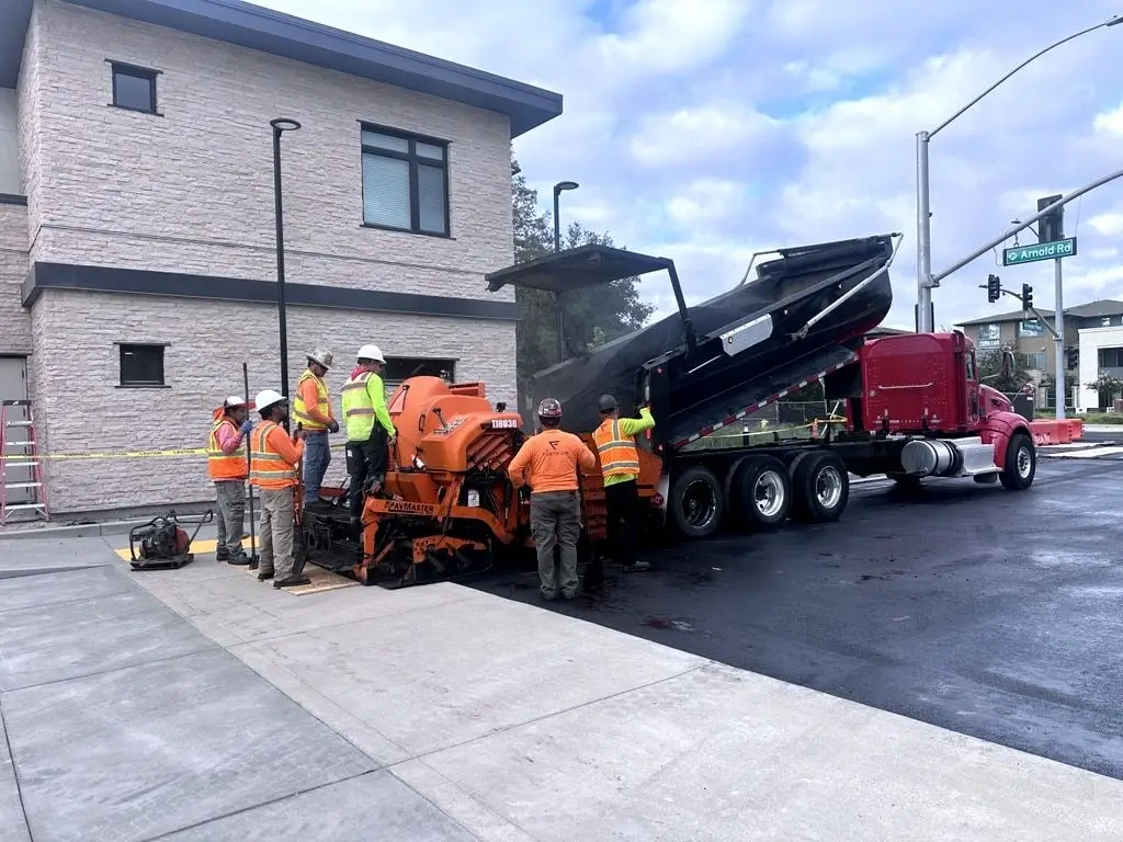 Crew members around the orange paver as a dump truck raises its bed to feed it, beside a new two-storey building.