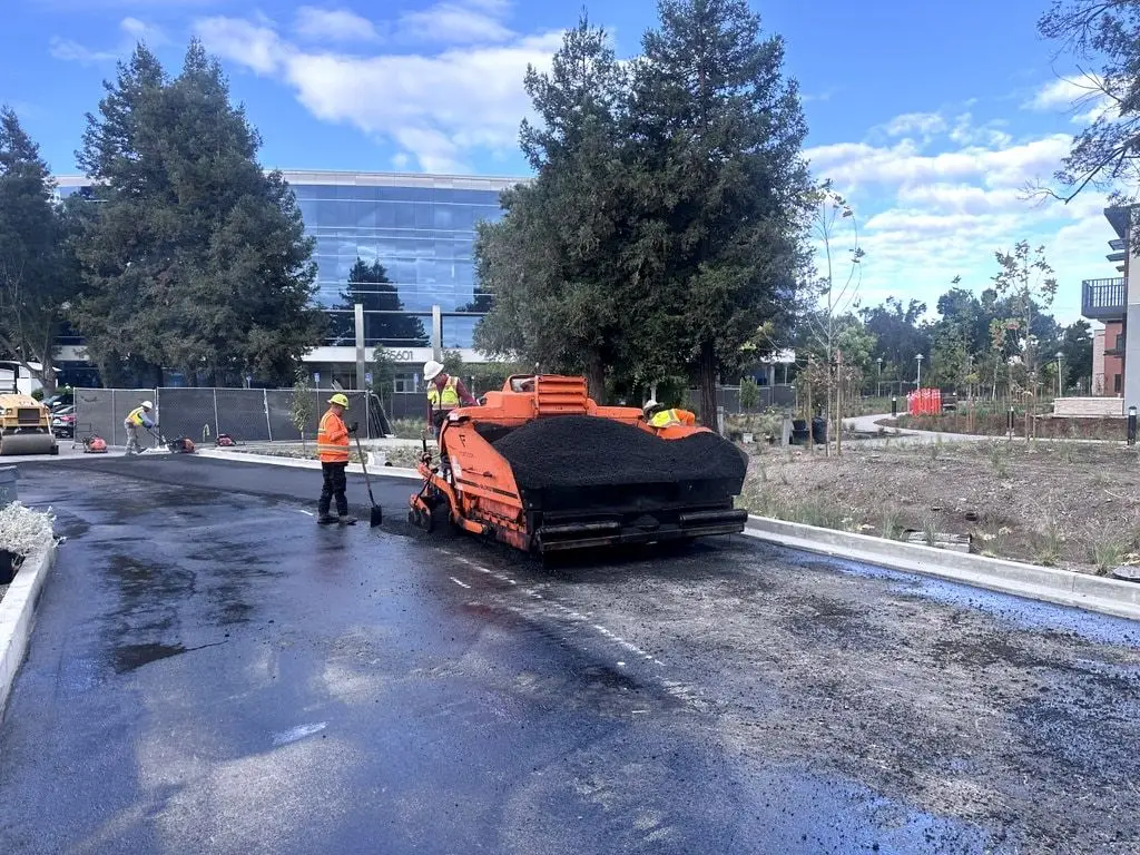 The paver placing hot asphalt along a new curb, with a glass office building behind the trees.