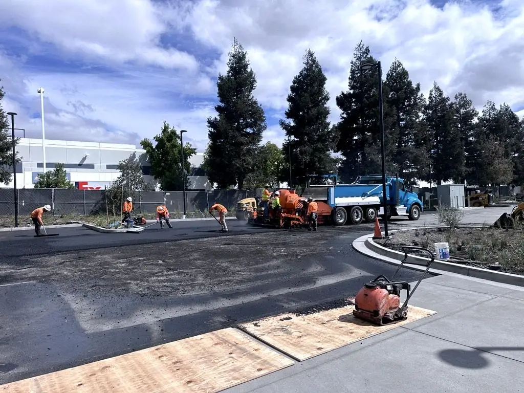 The paving crew at work beside a plate compactor in the foreground, with a blue dump truck behind the paver.