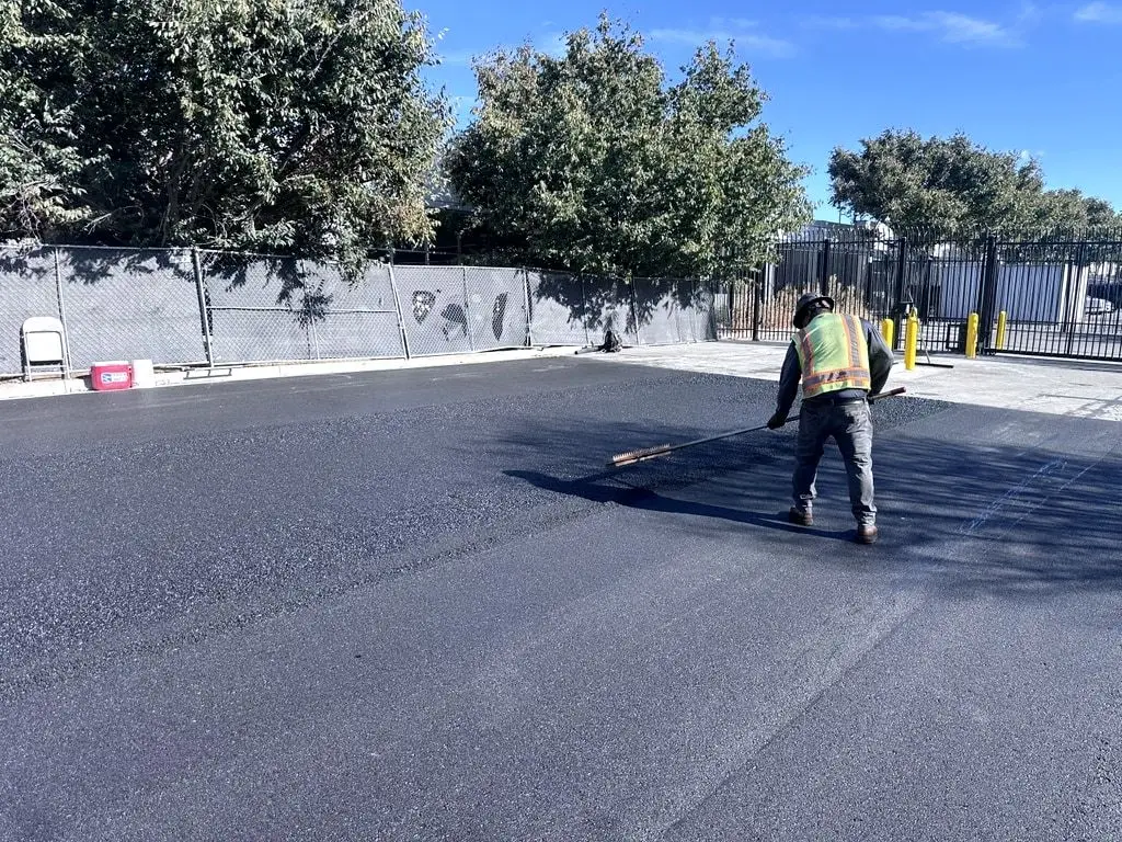 A worker raking the edge of fresh asphalt near a gate and bollards.