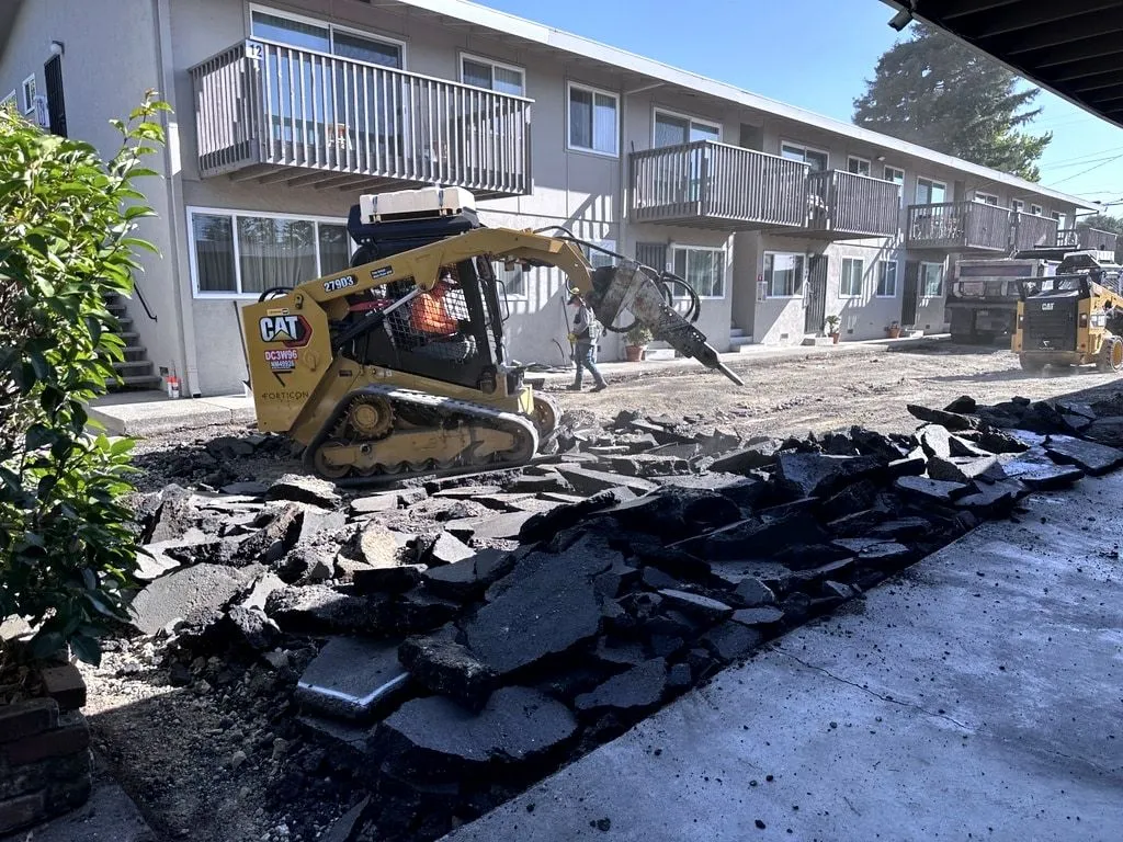 A skid steer with a breaker attachment breaking out old asphalt beside an apartment building, with the broken slabs piled along the edge.
