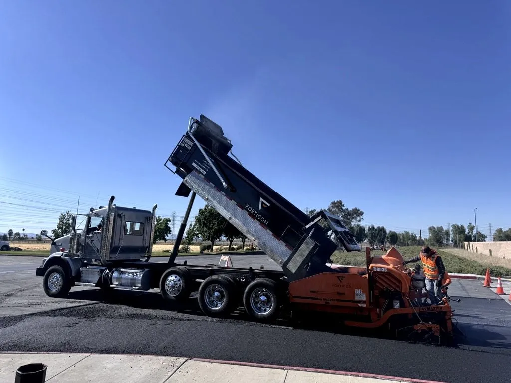 A Forticon dump truck with its bed raised, feeding hot-mix asphalt to the paver as the crew lays a new mat.