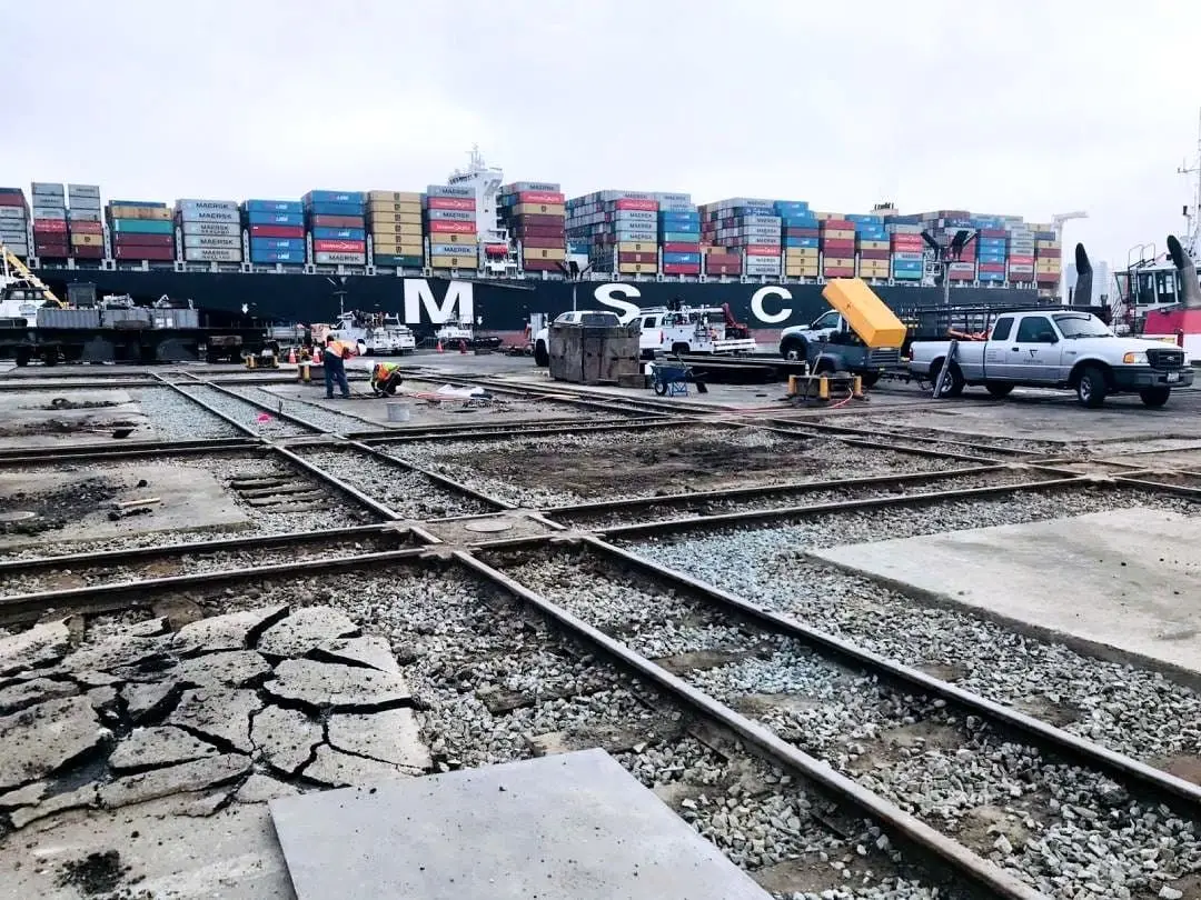 Broken pavement and embedded rail track at the Bay Ship Co. yard, with shipping containers stacked behind.
