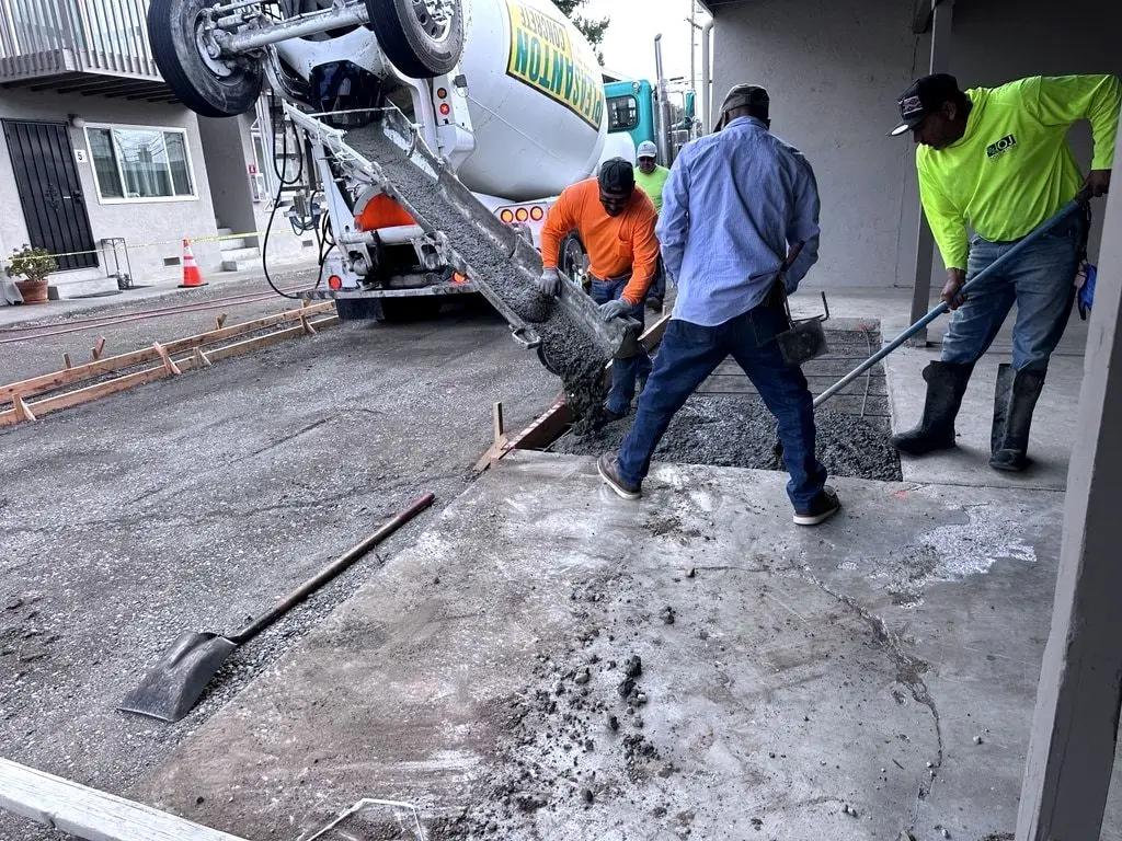 A mixer truck chute pouring concrete into forms while three workers spread it by hand.