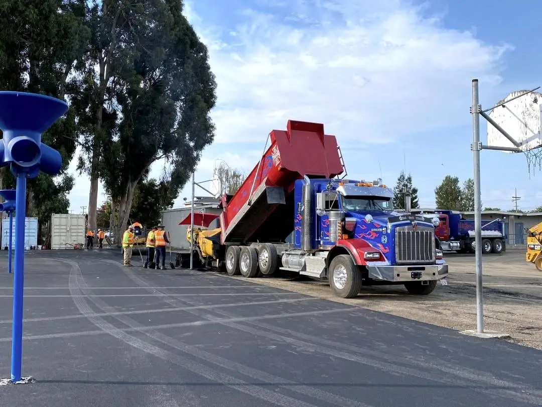 A dump truck with its bed raised, feeding hot-mix asphalt to the paver at Longwood Elementary School.
