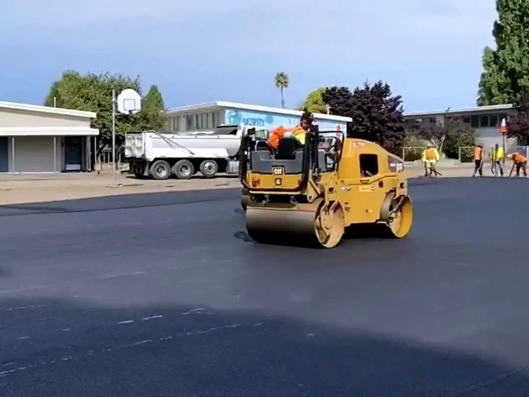 A roller compacting the new asphalt surface at Longwood Elementary School in Hayward, with the school buildings behind.