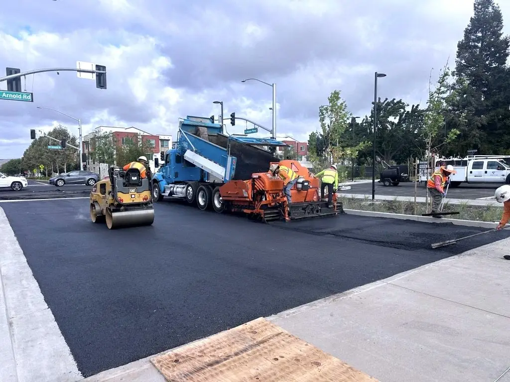 A roller and paver with a blue dump truck laying fresh black asphalt across a street intersection in Dublin, with new curbs and sidewalks beside it.