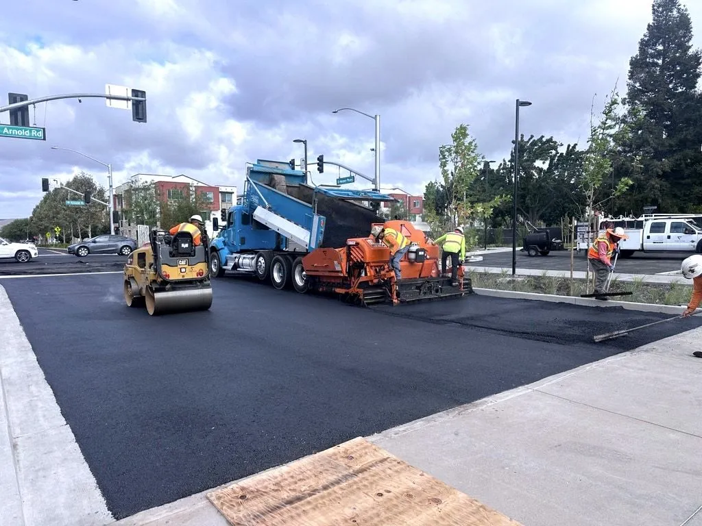 A roller and paver with a blue dump truck laying fresh black asphalt across a street intersection in Dublin, with new curbs and sidewalks beside it.