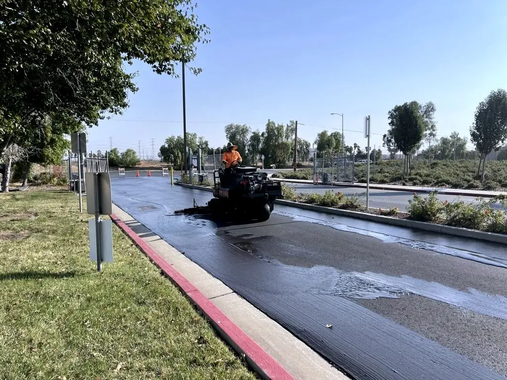 A crew member on a ride-on machine spraying fresh seal coat along a curb in a commercial parking lot.