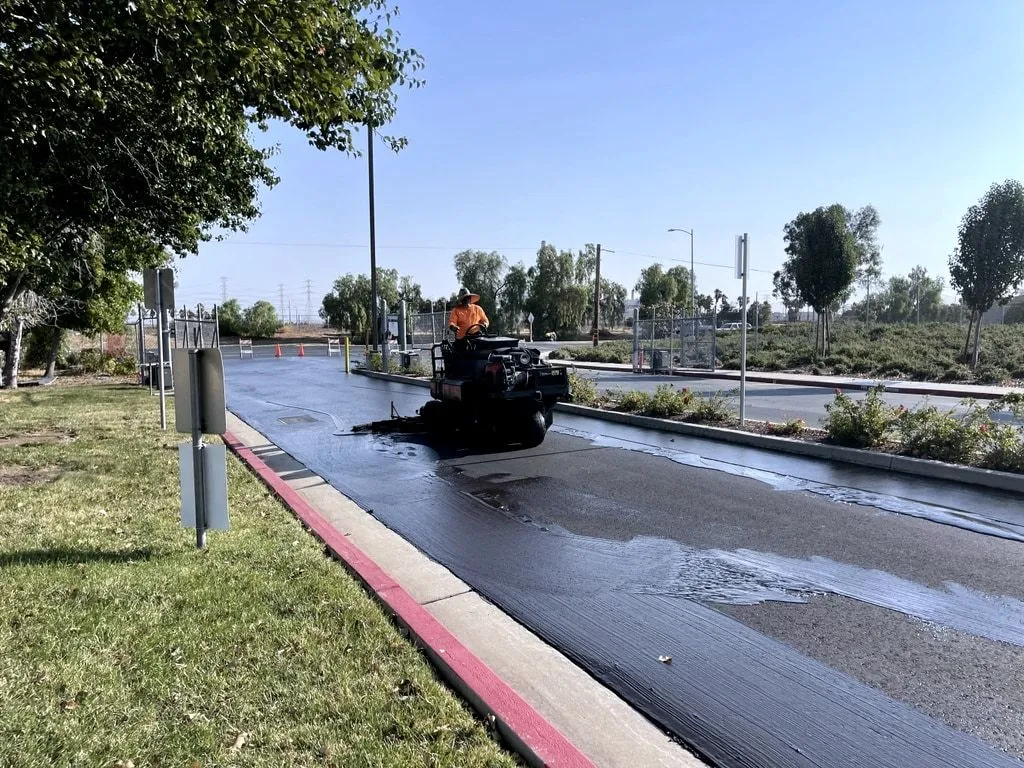 A crew member on a ride-on machine spraying fresh seal coat along a curb in a commercial parking lot.