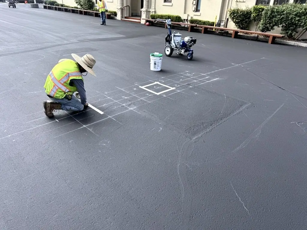 A crew member in a straw hat and hi-vis vest measuring and chalking the court layout on the sealed yard, with a striping machine behind.