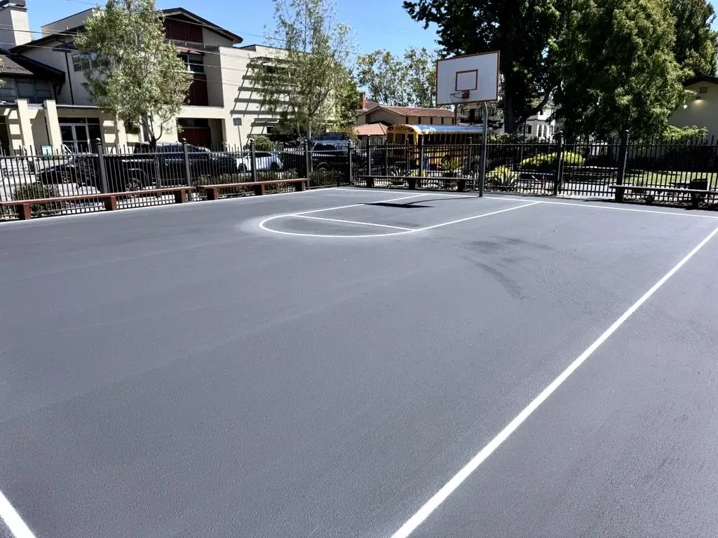 A finished basketball key and court line on the sealed yard, with a basketball hoop and a school bus beyond the fence.