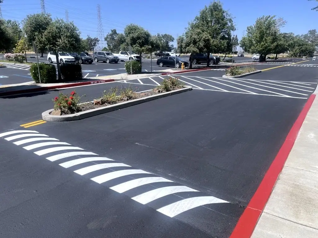 A freshly striped hatched crosswalk and stall lines on new asphalt, with a red-painted curb beside a landscaped island.