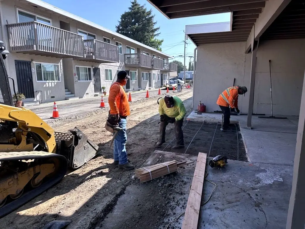 Crew members placing rebar in forms for the new concrete, with a skid steer beside them and apartments behind.