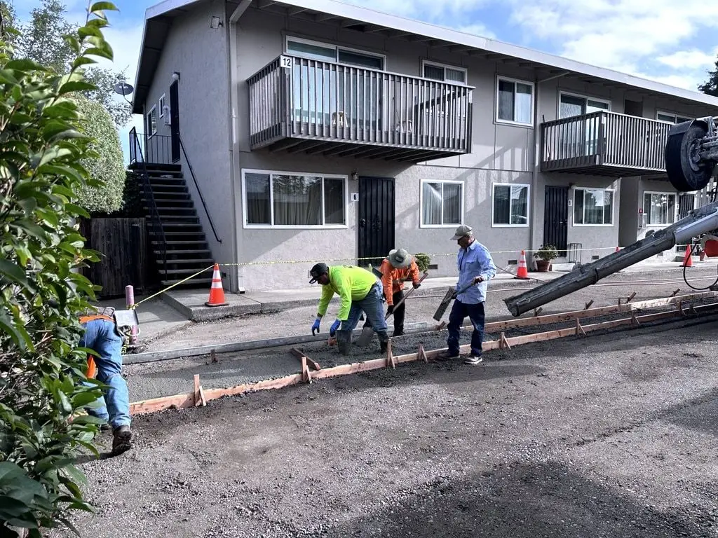 Crew members spreading and screeding concrete from a mixer chute into the forms in front of an apartment building.