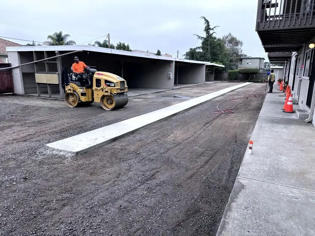 A roller compacting the graded base beside the new concrete gutter, with orange cones along the building.